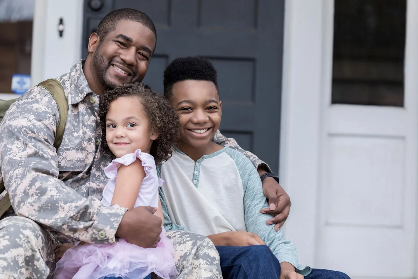 A man in a military uniform reunites with his two young children, who hug him in front of their home